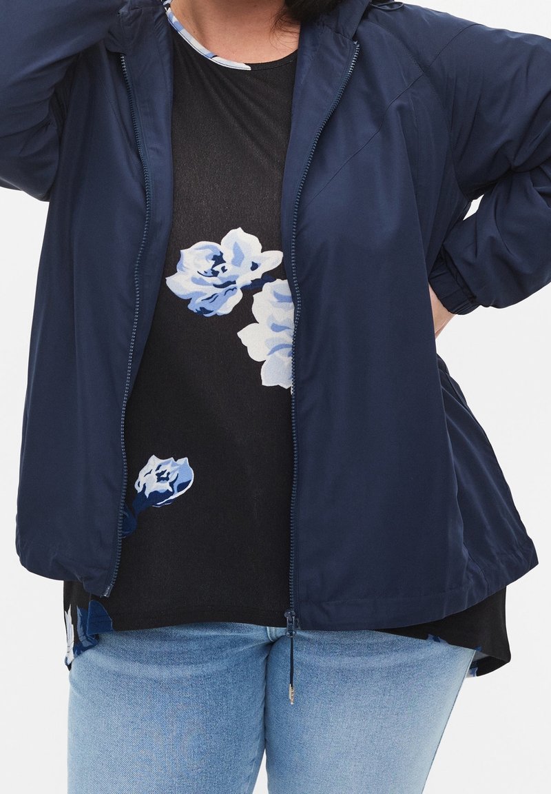 Kvinna klädd i en marinblå zip-up-jacka över en svart blommig skjorta, ihop med ljusblå jeans, stående med en hand på höften.