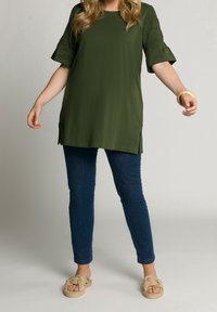 Olivgrön tunika med korta ärmar, med texturerade detaljer och sidoklyftor i nederkant. Matchad med mörkblå jeans och beige knutna sandaler.