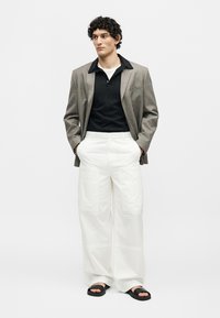 Homme portant un blazer marron à carreaux sur une chemise noire, un pantalon blanc à jambes larges et des sandales noires à bouts ouverts, debout les mains dans les poches.
