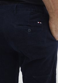 Pantaloni in corduroy blu navy con una tasca posteriore dotata di chiusura a bottone e un piccolo logo tricolore. Il tessuto ha un aspetto morbido e texture.