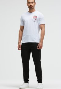 Weißes Baumwoll-T-Shirt mit rotem Aufdruck "CAMP DAVID PLAYER 01" auf der Brust, kombiniert mit schwarzen Jogginghosen und weißen Sneakern.