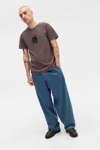 Brown graphic t-shirt featuring a central black logo and flames, paired with loose blue denim pants and black shoes, on a neutral background.