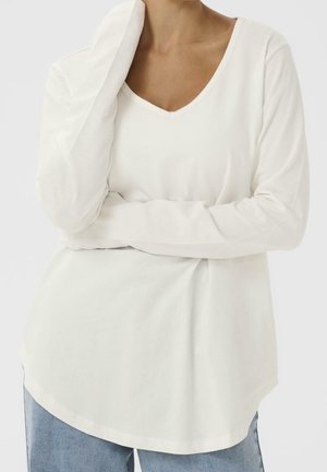 Personne portant un t-shirt blanc à manches longues avec un col en V et un jean bleu, un bras plié et la main touchant le cou.