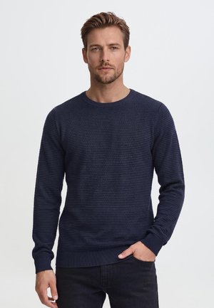 Homme aux cheveux bruns courts et à la barbe, portant un pull bleu marine texturé à manches longues et un pantalon noir, debout avec une main dans la poche.