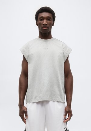 Férfi, aki ujjatlan szürke Nike pólót és fehér Nike rövidnadrágot visel, semleges háttér előtt áll.