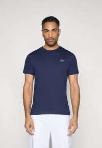 Granatowy t-shirt wykonany z bawełny, z okrągłym dekoltem i małym zielonym logo krokodyla na lewej stronie klatki piersiowej. Krótkie rękawy.