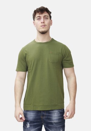 Giovane uomo con capelli corti e ricci che indossa una t-shirt verde oliva a maniche corte con tasca sul petto e jeans blu strappati, in piedi davanti a uno sfondo bianco.