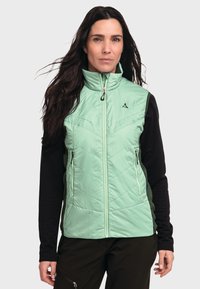 Frau mit langen, dunklen Haaren, die eine hellgrüne zip-up Weste über einem schwarzen Langarmshirt und dunklen Hosen trägt, steht vor einem schlichten Hintergrund.