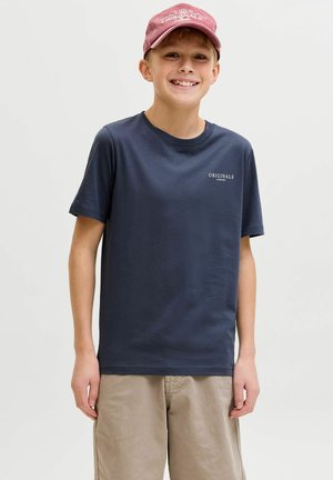 Niño sonriente con gorra roja, camiseta azul marino con la palabra "Originals" y pantalones cortos beige, de pie contra un fondo claro y sencillo.