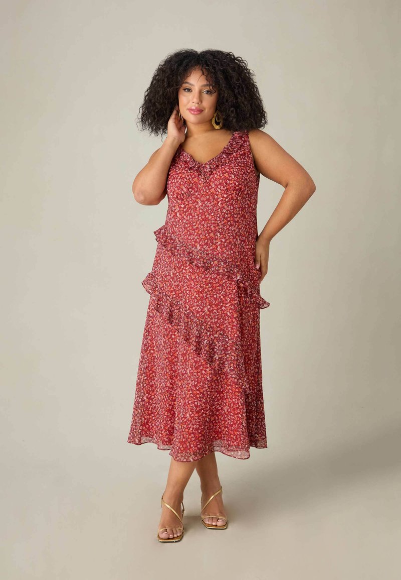 Röd blommig maxiklänning med volanglager och en V-ringning, gjord av lätt tyg; modellen är tillsammans med sandaler med remmar i guld.