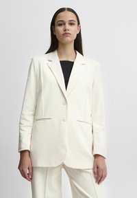 Slonovinový blazer vyrobený z měkké látky, s klopovým límcem, dvěma knoflíky a bočními kapsami. Design je přizpůsoben s uvolněným střihem.