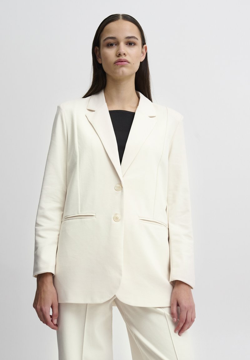 Slonovinový blazer vyrobený z měkké látky, s klopovým límcem, dvěma knoflíky a bočními kapsami. Design je přizpůsoben s uvolněným střihem.