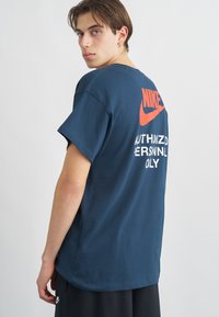 Μπλε βαμβακερό t-shirt με μεγάλο πορτοκαλί λογότυπο Nike και λευκό κείμενο στο πίσω μέρος, με πεσμένους ώμους και άνετη εφαρμογή.