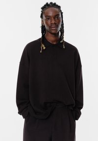 Zwart poloshirt met lange mouwen en een relaxte pasvorm, voorzien van een drievoudige knoopsluiting en een zachte textuur, gecombineerd met zwarte broek.