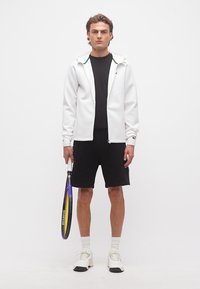 Άνδρας στέκεται κρατώντας ρακέτα τένις, φοράει λευκό φούτερ με φερμουάρ, μαύρο μπλουζάκι, μαύρο σορτς, λευκές κάλτσες και λευκά αθλητικά σε απλό φόντο.