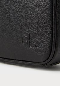 Black textured leather bag corner with embossed "cK" logo and smooth rounded piping on a white surface.