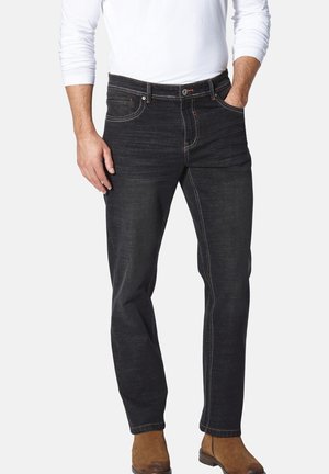 Man wearing dark denim jeans, white long-sleeve shirt, and brown suede ankle boots, standing with one hand in pocket.