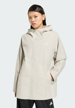 Giovane donna che indossa una giacca impermeabile beige chiaro con cappuccio e pantaloni neri con loghi Adidas, in piedi con una mano nella tasca della giacca.