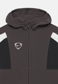 Σκούρο γκρι μπουφάν με κουκούλα και φερμουάρ στο μπροστινό μέρος, μαύρες πλαϊνές επιφάνειες, λευκές ρίγες στους ώμους και λευκό λογότυπο Nike στο αριστερό μέρος του στήθους.