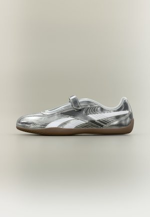 Baskets slip-on métalliques argentées avec des bandes blanches sur les côtés, une seule sangle Velcro, et une semelle en caoutchouc marron sur un fond neutre.