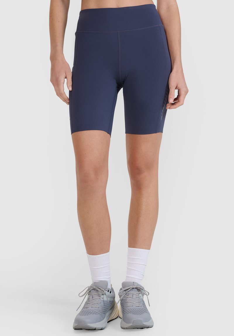 Femme portant un short bleu marine ajusté à hauteur des genoux, des chaussettes blanches et des chaussures de course grises, debout devant un fond uni.