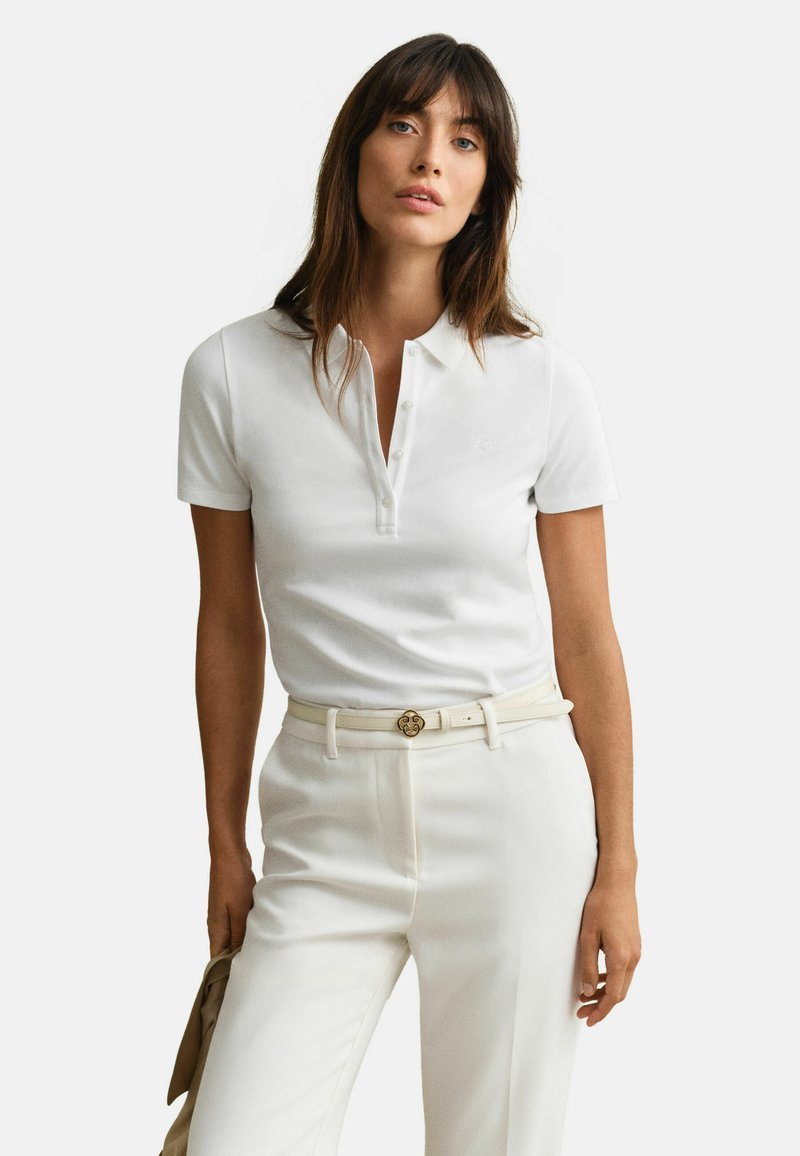 Femme aux cheveux longs bruns portant un polo blanc à manches courtes rentré dans un pantalon blanc taille haute avec une ceinture beige.