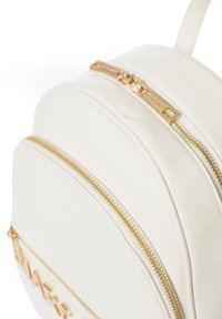 Sac à dos en simili cuir blanc avec des zips et des éléments en or. Dispose de deux compartiments zippés et d'un logo embossé sur le devant. Design arrondi.