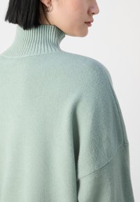 Világos mentazöld kötött pulóver magas bordás gallérral és oversized kialakítással, amely puha textúrával és esősen lelógó vállvarrásokkal rendelkezik.