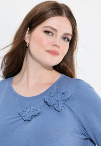Sininen lyhythihainen paita pyöreällä pääntiellä, jossa on kaksi samasta kankaasta valmistettua tekstuurilla varustettua kukkakoristetta rinnassa. Pehmeä ja kevyt materiaali.