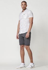 Camisa blanca de manga corta con estampado de hojas de palma, diseño con botones en la parte delantera, bolsillo en el pecho; combinada con pantalones cortos grises y zapatillas blancas.