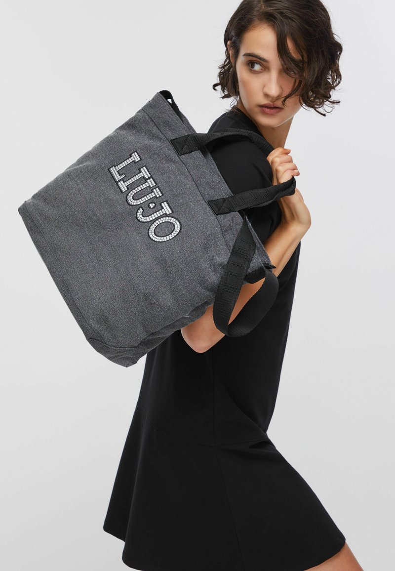 Femme en robe noire portant un grand sac cabas gris avec le logo "LIUJO" sur l'épaule, sur un fond clair uni.