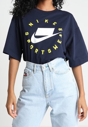 Osoba nosząca granatowy t-shirt Nike Sportswear z żółtym napisem i jasnoniebieskie jeansy z wysokim stanem na jednolitym tle.