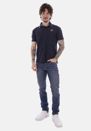 Uomo con capelli ricci e tatuaggi che indossa una polo blu navy, jeans blu e sneakers bianche, in piedi con una mano in tasca.