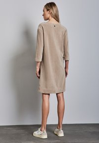 Robe en velours côtelé beige avec des manches trois-quarts, un col rond et un petit détail de bouton à l'arrière, portée avec des baskets beiges.