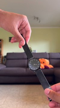 Reloj de pulsera negro con una esfera redonda, correa de malla metálica, esfera minimalista y acentos plateados. Una mano sostiene el reloj contra un sofá.