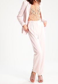 Lichtroze op maat gemaakte blazer over een kantblouse, gecombineerd met bijpassende geplooide broek en blush enkelsandalen. Soepele textuur en strakke pasvorm.