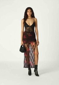Jovem mulher num vestido pret-a-porter preto e transparente com padrão floral, segurando uma pequena mala preta, vestindo botas pretas até ao tornozelo, em pé contra um fundo simples.