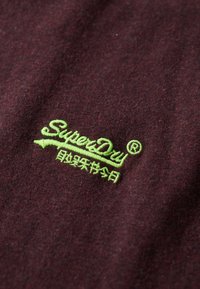 Burgundowa tkanina z wyraźnym, zielonym haftowanym logo "SuperDry" oraz zakrzywionym akcentem pod tekstem. Teksturowana powierzchnia, casualowy design.