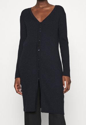 Personne portant un long cardigan noir boutonné avec un col en V sur un pantalon noir, devant un fond blanc uni.