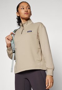 Beige fleece pullover met halve rits, voorzien van een logopatch, geribbelde mouwen en zijzakken, gedragen over donkere leggings, met een lichtblauwe strap.