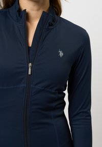 Vrouw in een aansluitende marineblauwe zip-up jas met een klein geborduurd polo speler logo op de borst, rits gedeeltelijk open.