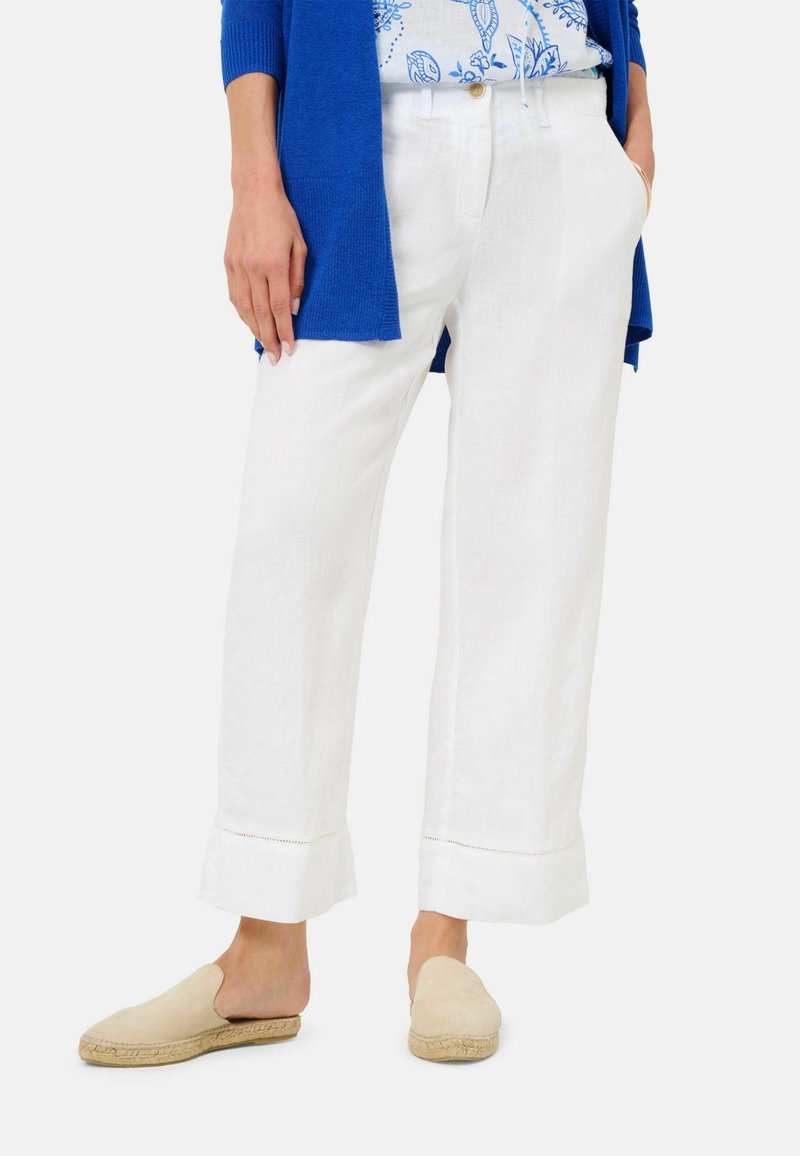 Femme portant un pantalon en lin blanc à jambes larges et coupés, des espadrilles beiges à enfiler et un cardigan bleu avec un top fleuri, les mains dans les poches.