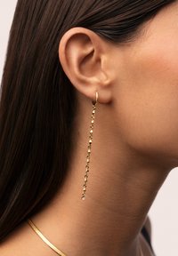 Pendiente de aro dorado con una cadena larga que presenta pequeños eslabones triangulares y un único acento de cristal, exhibido sobre un fondo neutro.