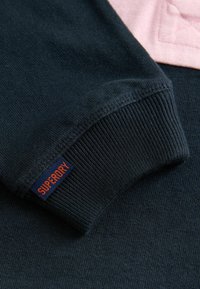 Navy Sweatshirt mit gerippten Bündchen und einem orangen "SUPERDRY"-Label. Der Stoff hat eine weiche Textur, die durch einen hellen rosa Akzent ergänzt wird.