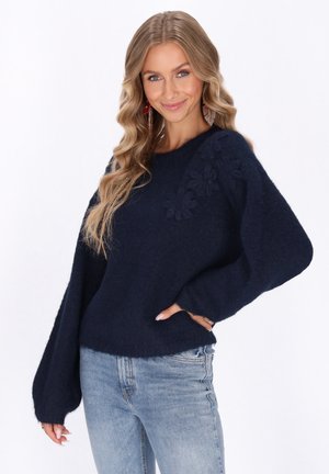 Navyblauwe gebreide trui met textuur van bloemaccenten op de schouder, brede mouwen en een ontspannen pasvorm. Gecombineerd met lichtblauwe jeans.