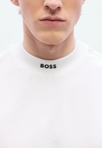 Λευκό t-shirt με στρογγυλή λαιμόκοψη που φέρει μαύρο λογότυπο "BOSS" στο γιακά. Το υλικό φαίνεται λείο και απ αλό, με απλό και καθαρό σχεδιασμό.
