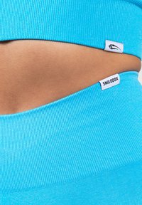 Blauer, gerippter Stoff-Crop-Top, das Haut darunter zeigt, mit zwei Etiketten: eines mit einem Logo und das andere mit "SMILODOX." Glatte Textur.