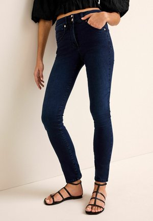 Donkerblauwe denim skinny jeans met een hoge taille, met voorzakken en metalen knopen. Gekleed met zwarte sandaaltjes met bandjes.