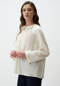 Blusa larga e a maniche lunghe in tessuto bianco sporco con scollo rotondo e dettagli arricciati al collo. Tessuto liscio e vestibilità comoda.