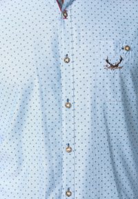 Světle modrá košile s malými tmavými křížovými vzory, hnědými knoflíky, kapsou na prsou a vyšitými parohy jelena s kurzívou.